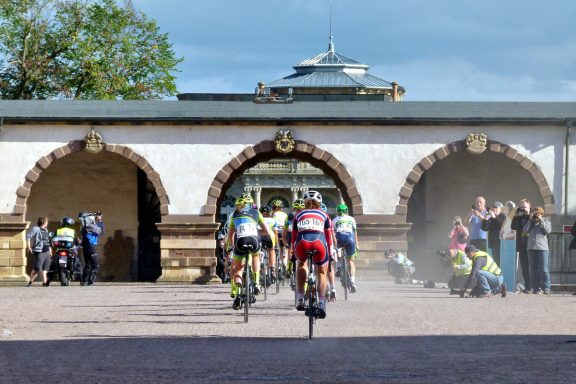 Radsportler fahren durch eine architektonische Struktur mit Zuschauern im Hintergrund.
