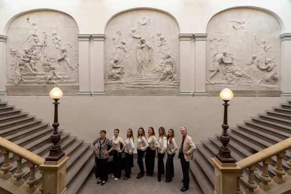 Gruppe von sieben Personen auf einer Treppe vor skulpturalen Wandreliefs.