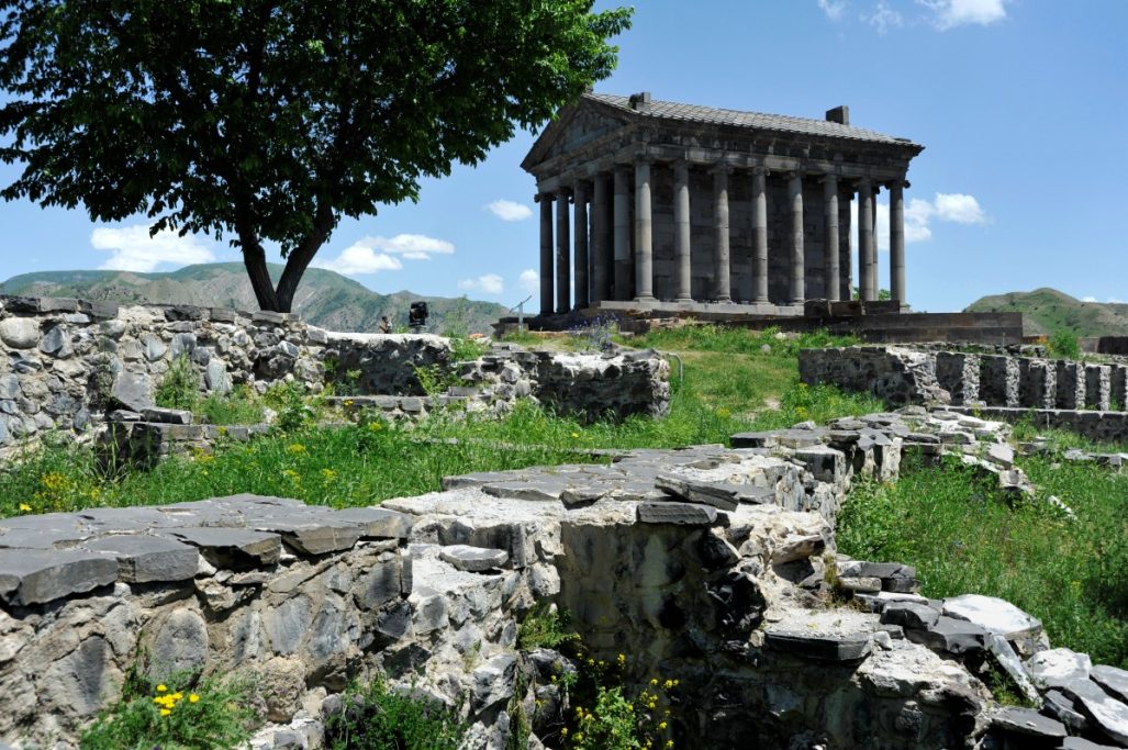 Römischer Tempel mit Ruinen und Bäumen in einer grünen Landschaft unter blauem Himmel.