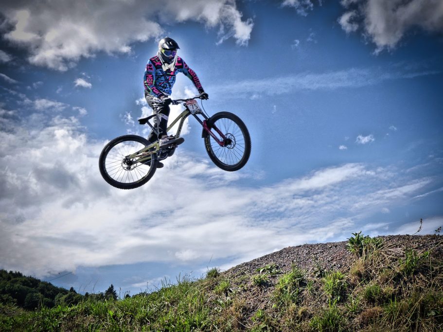 Fahrer springt mit Mountainbike über einen Hügel unter blauem Himmel.